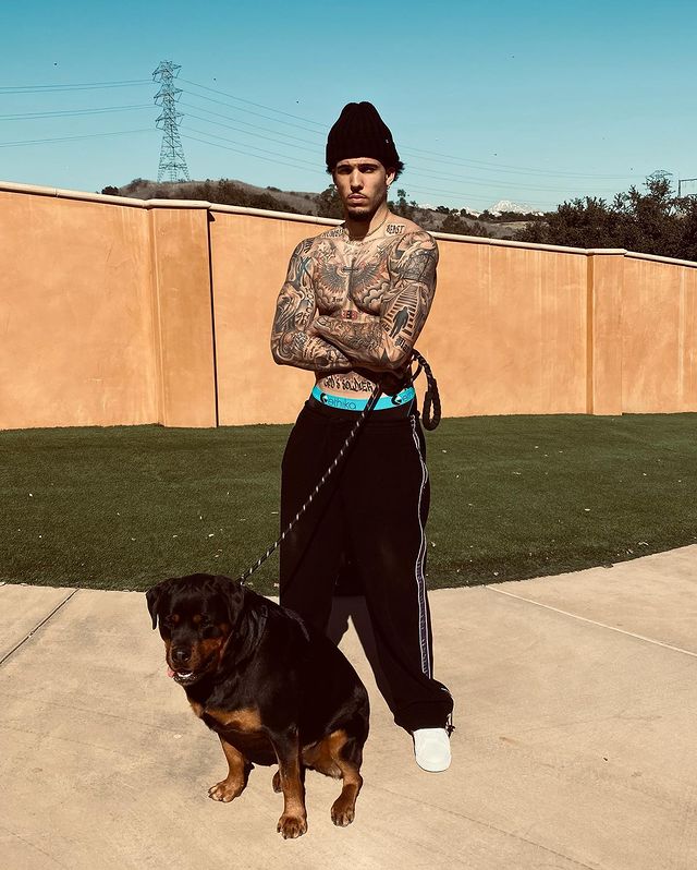 LiAngelo Ball showing his tattoo with his pet. He is wearing a black woolen cap, black sweat pant and white shoes.